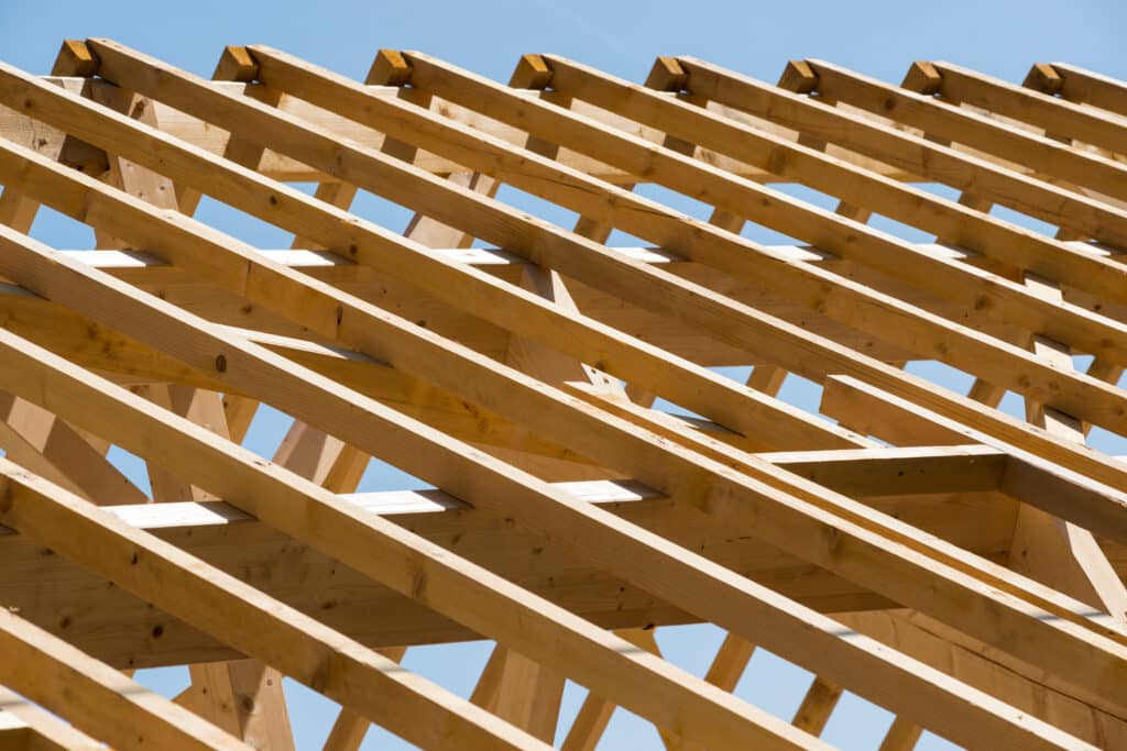 Charpente de toit en bois – Construction & Rénovation Charpente de toit en bois clair contre un ciel bleu lumineux, montrant la structure d'une maison en construction avec chevrons.