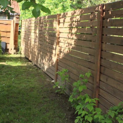 Clôture en bois horizontale dans un jardin ensoleillé avec abri et buissons verts en arrière-plan.