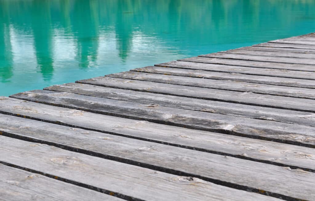 Lac turquoise et ponton en bois : Sérénité naturelle Ponton en bois gris patiné avec des planches marquées, s'avançant sur un lac d'eau turquoise claire et agitée par de légers reflets.