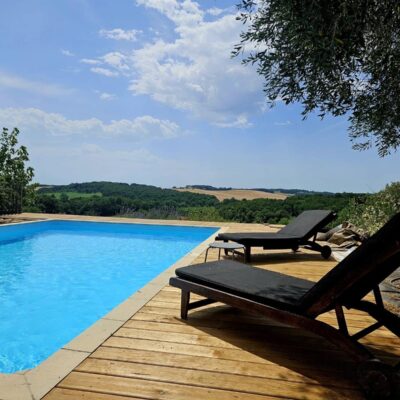 Piscine avec terrasse en bois, transats noirs et vue sur les collines verdoyantes sous un ciel bleu.