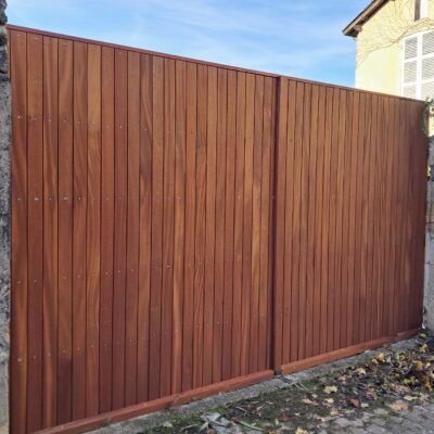 Portail moderne en bois exotique à lames verticales marron devant une maison ensoleillée.